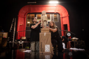 Two people kneel on a stage, bringing a wooden puppet to life atop stacked boxes, surrounded by theater props, equipment, and a large red arch. In the background, set pieces evoke both a standing tree and a fallen tree.